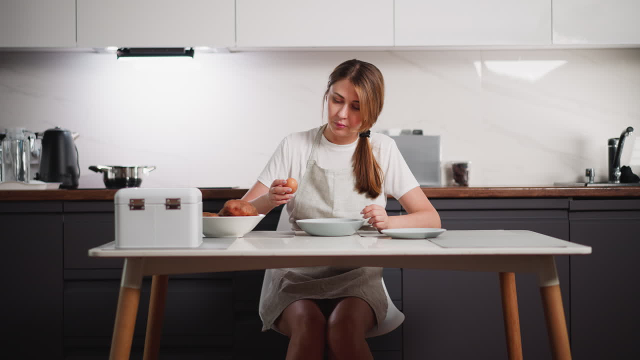 Young female caterer wearing apron breaking egg on glass plate while seated at kitchen table with bowls and utensils, surrounded by modern cooking appliances in well-lit domestic kitchen interior