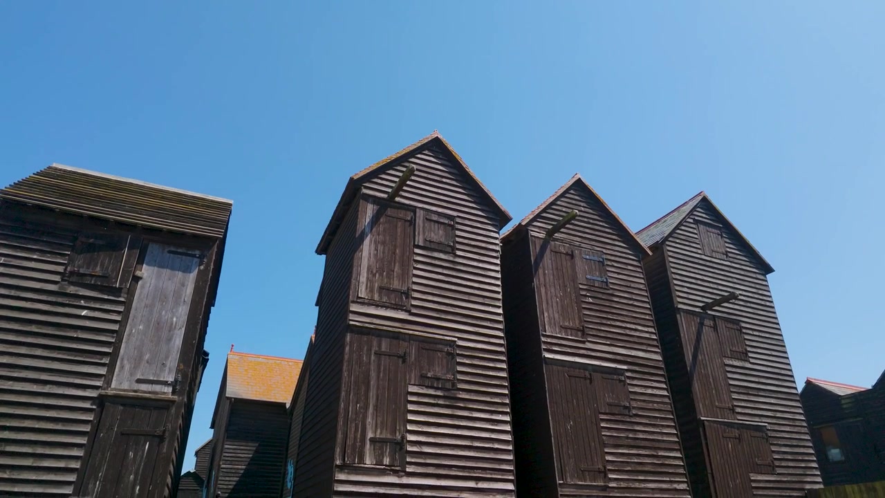 Les cabanes de pêcheurs de Hastings. La caméra descend d'un ciel bleu clair, avec un grand espace pour le texte, pour révéler les cabanes de pêcheurs en bois goudronné sur la plage de Hastings, en Angleterre