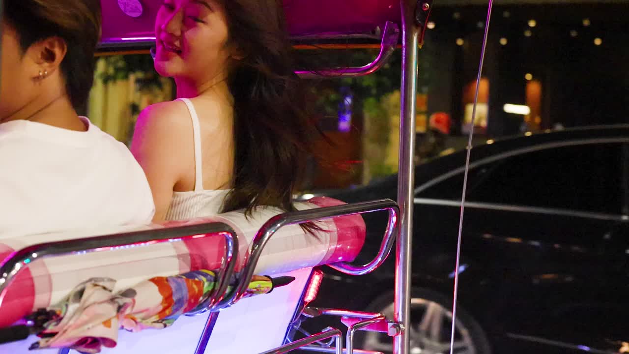 Una pareja sonriente viaja en un tuk-tuk abierto por una vibrante calle nocturna con colorida iluminación de neón.