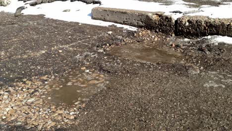 Moviéndose-A-Lo-Largo-De-Un-Camino-Roto-Con-Nieve-En-Los-Bordes-Y-Baches-Llenos-De-Agua-Y-Rocas