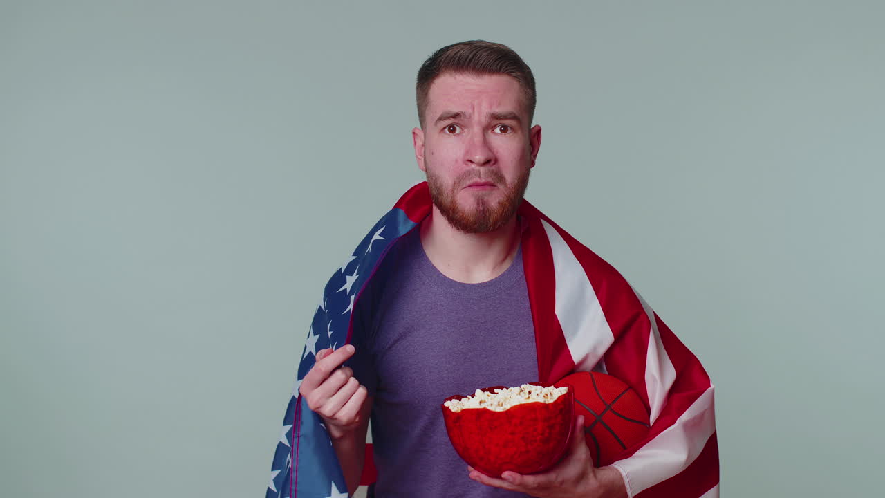 수염이 있는 젊은 남자 농구 팬이 팝콘을 먹고 우승 제스처를 하고 승리를 축하하고 있습니다.