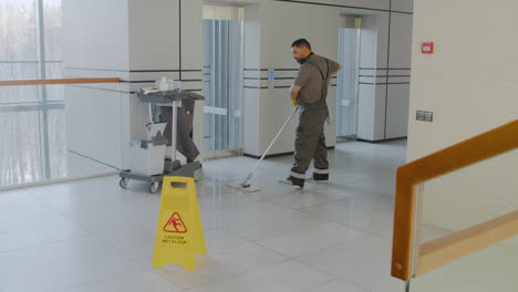 vista lejana del hombre de limpieza árabe limpiando con mope detrás de una señal de advertencia de piso húmedo