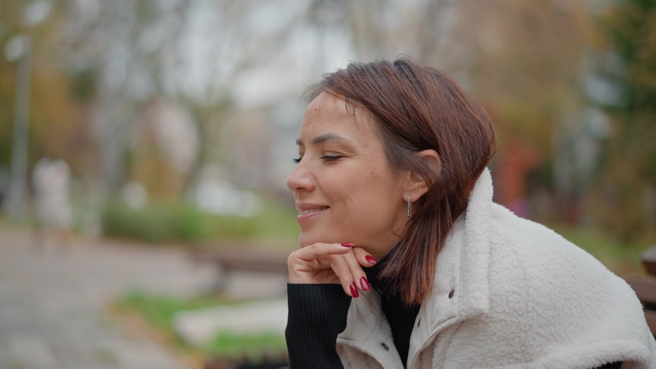 Middle-aged woman seated outdoors in autumn park, resting chin on hand, wearing casual vest and showing soft smile while looking outward beside blurred trees and pathway, with woman, middleaged