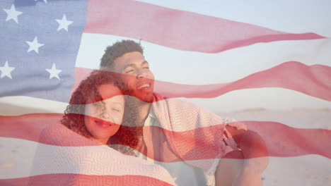 Animation-of-american-flag-over-happy-diverse-couple-sitting-on-sunny-beach-embracing