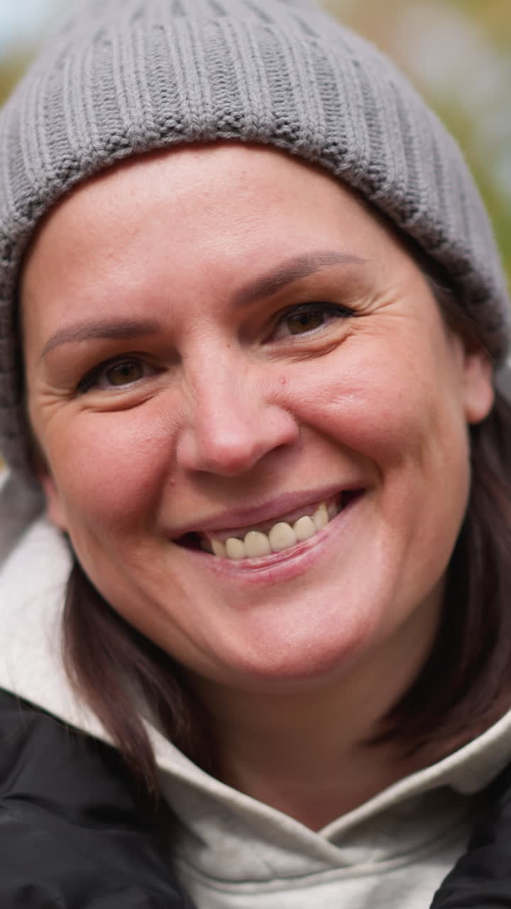 mujer con gorro de ceniza y chaqueta rosa soplando hojas secas de otoño de sus manos con una cálida sonrisa, uñas pintadas de negro, rodeada de colorido follaje borroso de otoño y árboles en el fondo