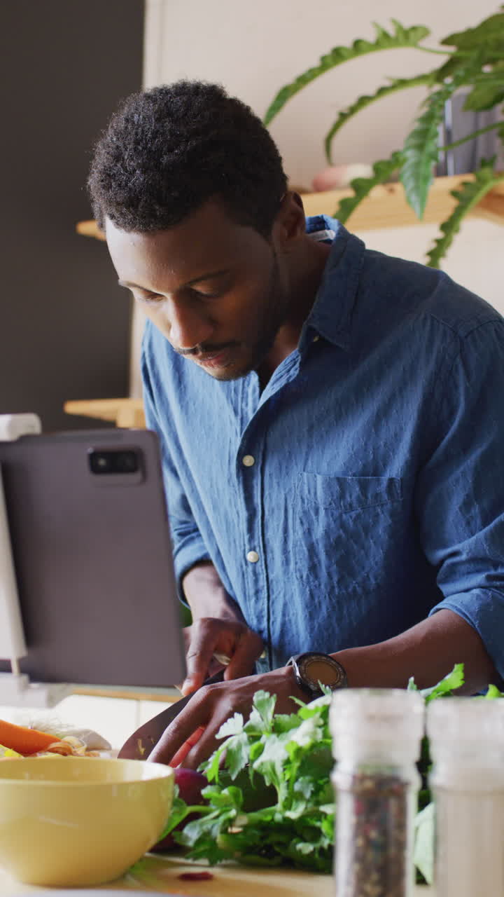 Vertical video of african american man cooking dinner in kitchen, using tablet