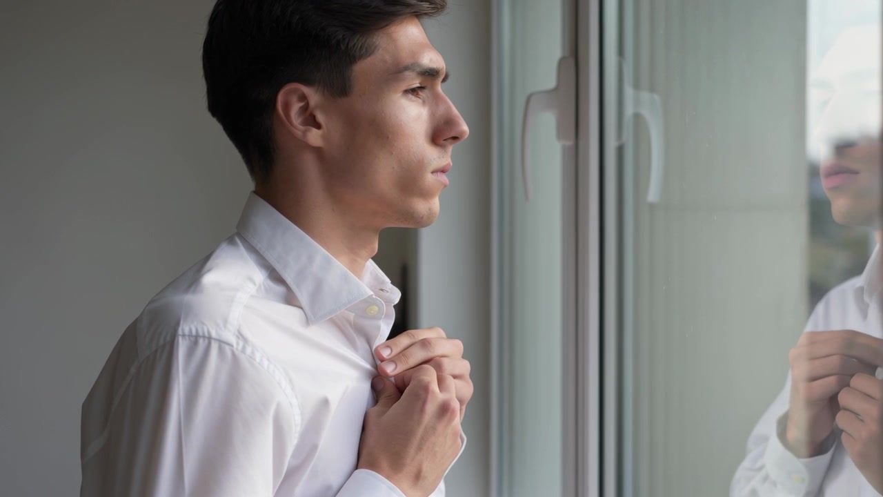 窓際で白いワイシャツを着た男性が、襟を整えながら自分の姿を確認している。