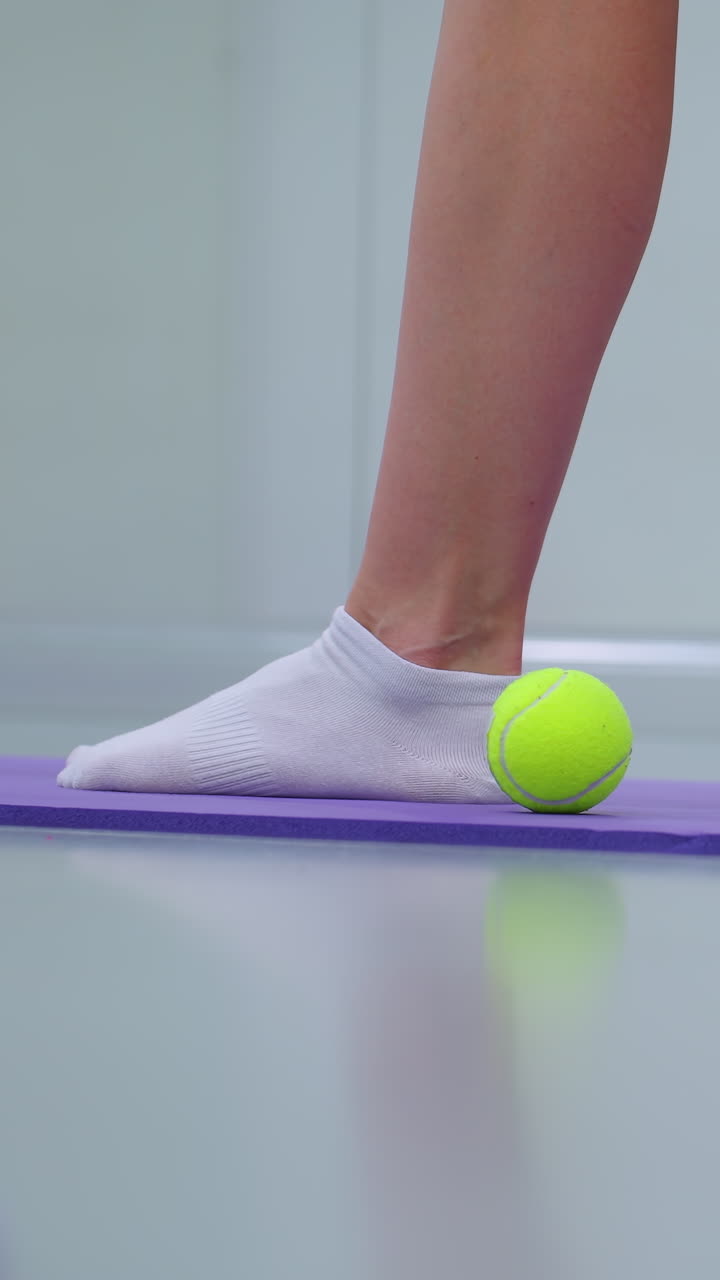 vista lateral de la pierna de un ejercicio de fitness en una alfombra púrpura con una pelota de tenis verde cerca, el espejo refleja a la persona estirándose hacia adelante, mostrando el equilibrio y el movimiento controlado en un entorno de gimnasio activo