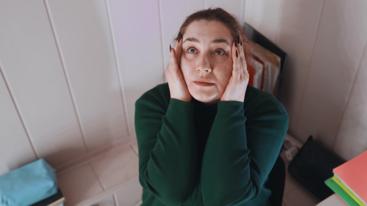 White woman seated at cluttered home desk with folders and notebooks, overhead and frontal angles show her massaging temples then pausing in reflection, quiet moment of breath, shifting