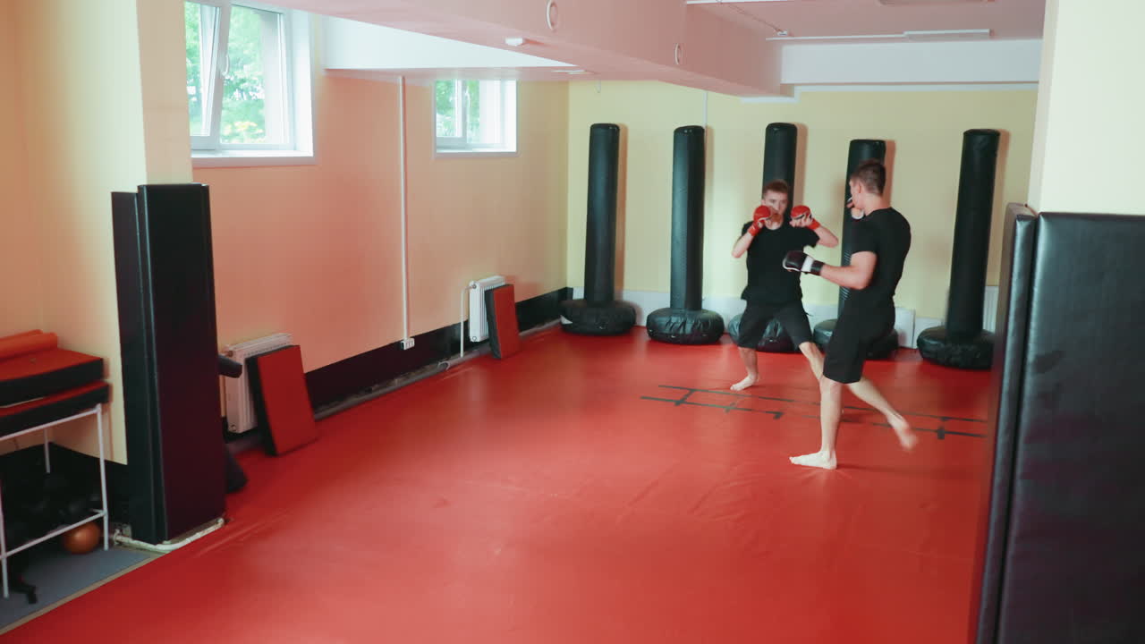 글러브를 착용하고 빨간색 매트 바닥에서 공격 및 방어 자세를 연습하며 펀칭 백으로 둘러싸인 무술 체육관에서 겨루는 가라테 수련생