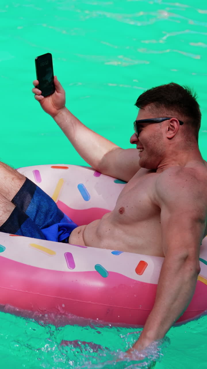 Muscular male sits in the inflatable circle holding a phone. Man taking selfie while resting in the pool. Vertical video