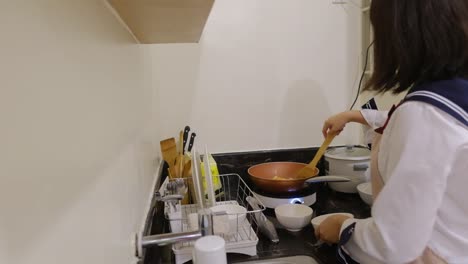 Medium-static-shot-of-a-young,-Asian-woman-stirring-and-cooking-food-in-pan-with-a-wooden-spoon