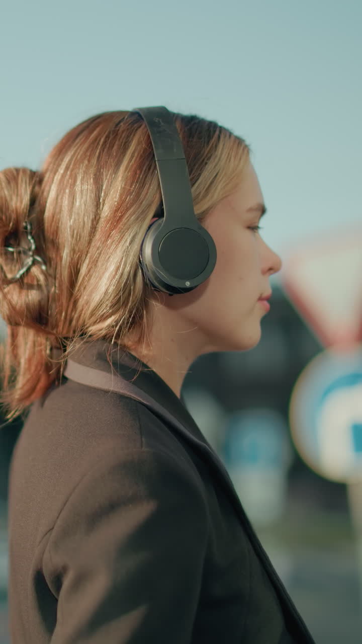 Back view of lady in black suit with messy bun enjoying music through headphones while holding coffee cup, walking under sunlight ambiance with urban background featuring buildings and parked cars