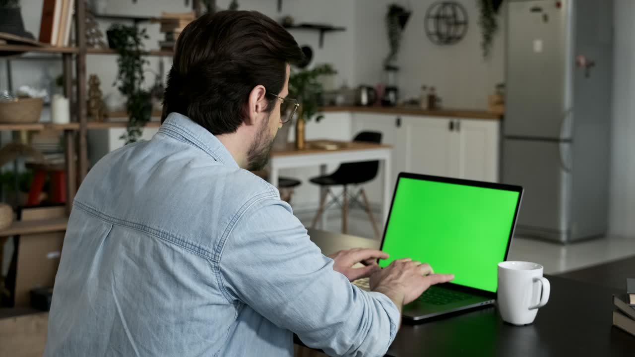 sobre el hombro de un joven caucásico sentado, trabajando desde casa en una computadora portátil, pantalla verde clave de croma