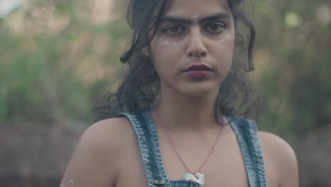 Slow-motion-handheld-shot-of-a-young-indian-model-dressed-in-a-denim-dress-and-a-necklace-with-a-horse-looking-flirtatiously-into-the-camera-and-standing-in-the-garden