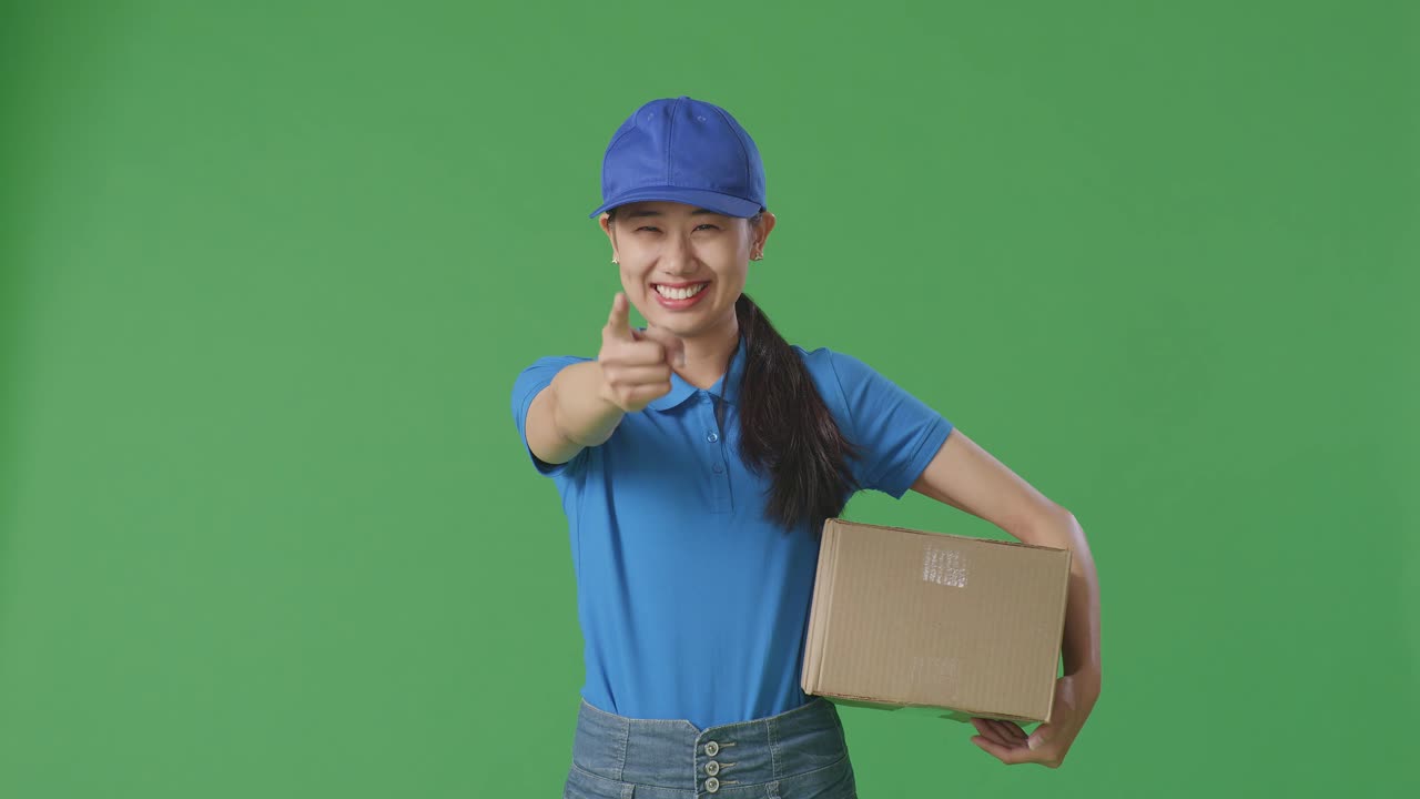 Corriera asiatica in uniforme blu che sorride e fa un gesto di onestà mentre consegna un pacco su sfondo green screen in studio
