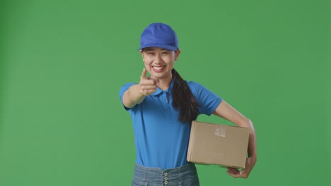 asian female courier in blue uniform smiling and making honest gesture while delivering a carton on green screen background in the studio