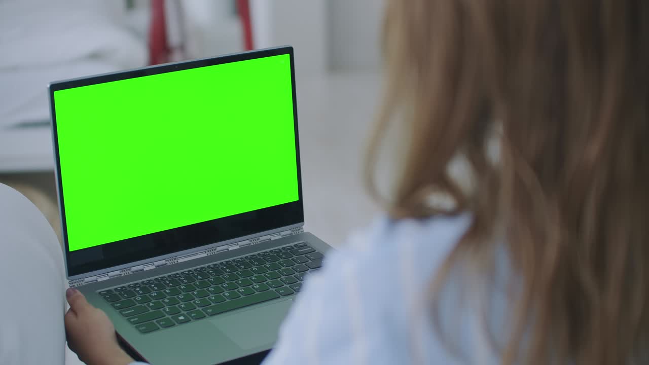 mujer joven en casa trabaja en una computadora portátil con pantalla de maqueta verde. ella sentada en un sofá en su acogedora sala de estar. sobre el hombro de la cámara disparado