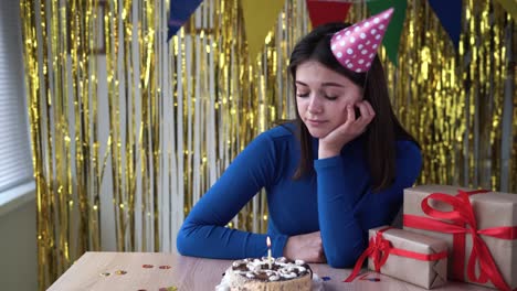 cumpleaños aburrido de una deprimida dama caucásica en casa sola. la chica está aburrida sentada en la mesa con un regalo en un sombrero festivo y pastel. decoraciones de vacaciones.