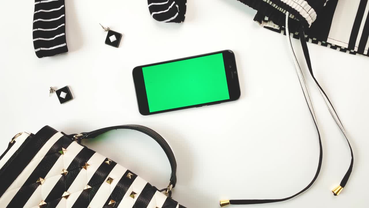Striped female hat, earring, necklace, black and white purse with stripes on white background. Smartphone with green screen. Flat lay, top view. Chroma key, mock up.