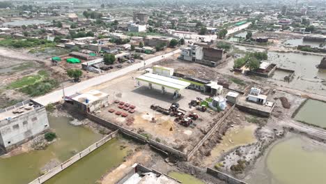 Aerial-circular-shot-of-a-fuel-pump-in-rural-Badin,-Sindh,-amidst-buildings-and-water