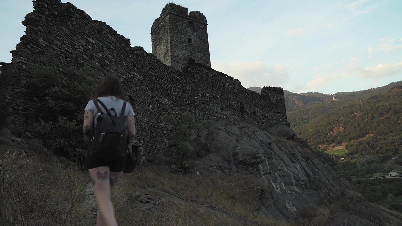 Smuk pige går på gaden for at nå middelalderborgen med sin rygsæk på en solrig dag i Valle d'Aosta, Italien