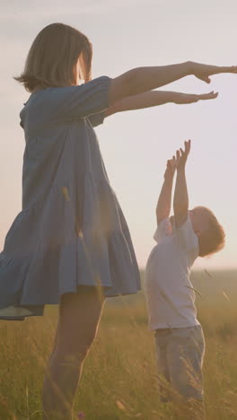 una tierna escena de una madre con un vestido azul que fluye levantando los brazos mientras su hijo pequeño, vestido con una camiseta blanca, se extiende ansiosamente para conocer sus manos. se paran en un campo de hierba pacífico y iluminado por el sol