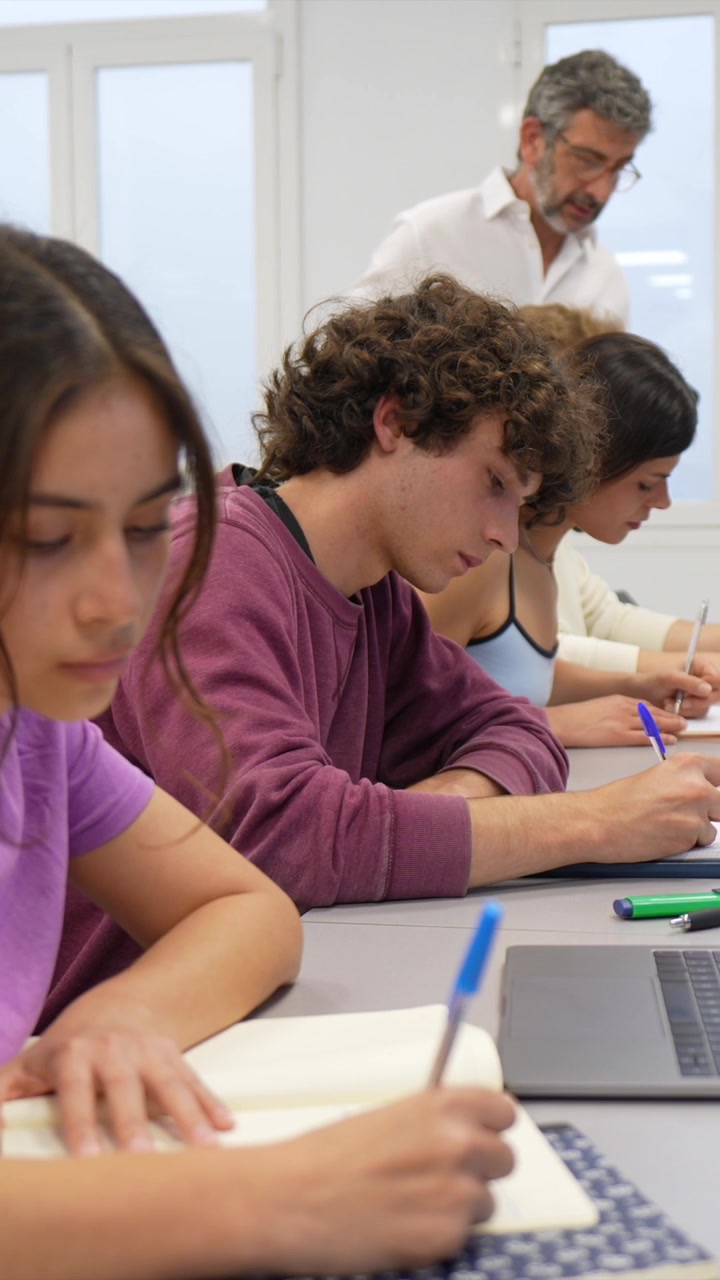 Profesor supervisando a estudiantes tomando apuntes en un aula moderna.