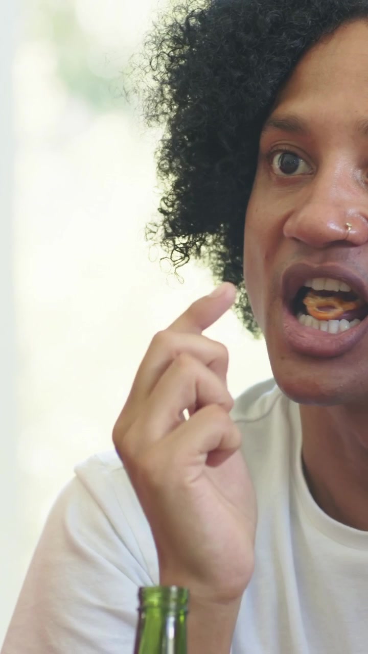 Video verticale: Uomo in maglietta bianca che porta un pretzel alle labbra e mangia vicino a una finestra, bottiglia verde, spazio per il testo