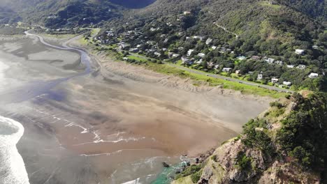 drone tilt-up next to lion rock reveals charming piha village, west auckland