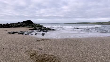 Footsteps-in-the-sand-and-black-shiny-rocks-with-gentle-waves-washing-the-shore-in-slow-motion