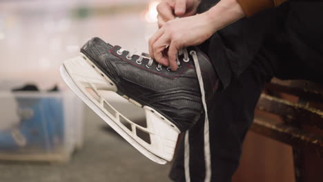 a close-up view of someone loosening the laces of a black ice skate with two hands, with a blue skating shoe visible in a transparent container in the background