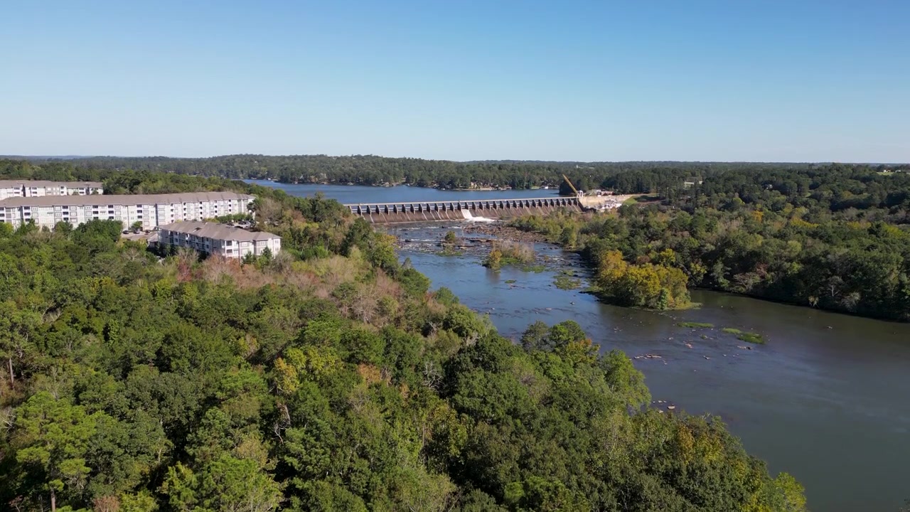 Přehrada Oliver na řece Chattahoochee provozovaná společností Georgia Power se čtyřmi turbínami a kapacitou šedesát megawattů, tvořící nádrž Lake Oliver, jak se stavba tyčí nad hustým lesem, letecký záběr z dronu stoupajícího vzhůru.