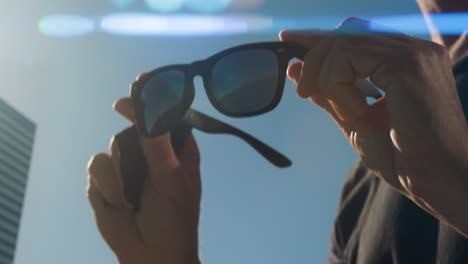 cerca de las manos pulindo la lente derecha de gafas de sol negras, hombre caucásico con camiseta negra, cielo azul y edificio en el fondo, llamaradas anamórficas de la luz solar