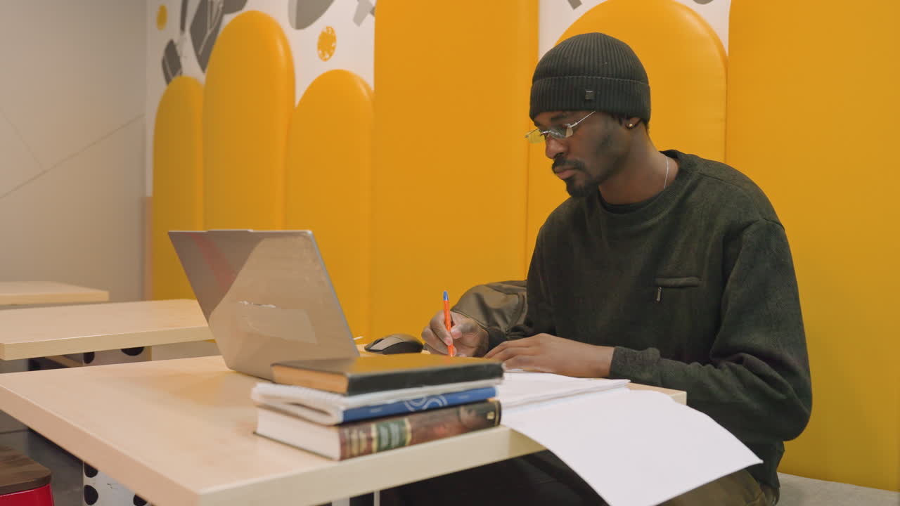 Ung mann studerer ved laptop i gul studiebod, blar i sider og skriver notater, sjonglerer lærebøker mens han forbereder seg til eksamen, skisserer ideer til frilans designprosjekt, undersøker referanser