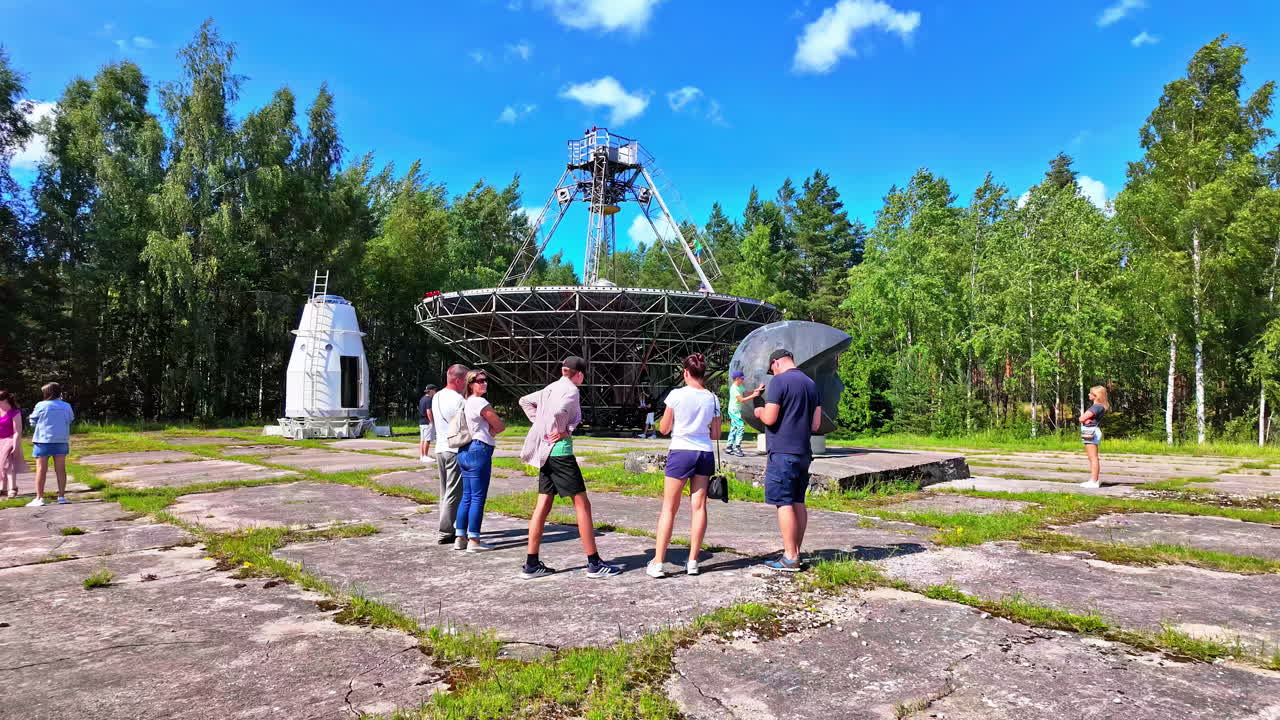People observe a large radio telescope in a forest under a clear blue sky
