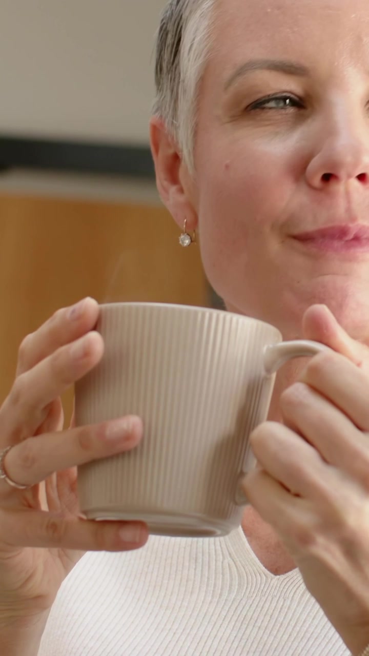 Video verticale: Donna matura con top bianco che porta la tazza alla bocca, sorseggiando una bevanda a casa e mostrando un anello.