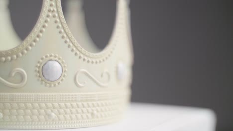 close up of a beige crown on a pedestal with ornaments and white pearl jewels with peaks jagged out of plastic. with a neutral grey background. smooth backwards dolly camera movement. shot in 4k.