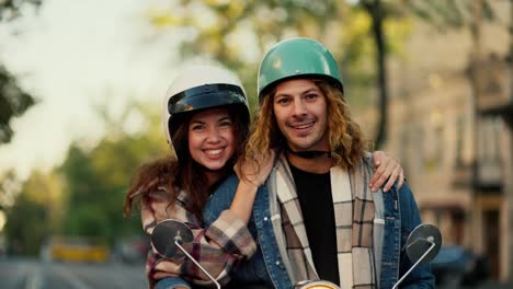 portrait of a happy couple, a brunette girl in a plaid shirt hugs her boyfriend with long curly hair in a denim jacket and a green motorcycle helmet near her motorcycle in the summer city