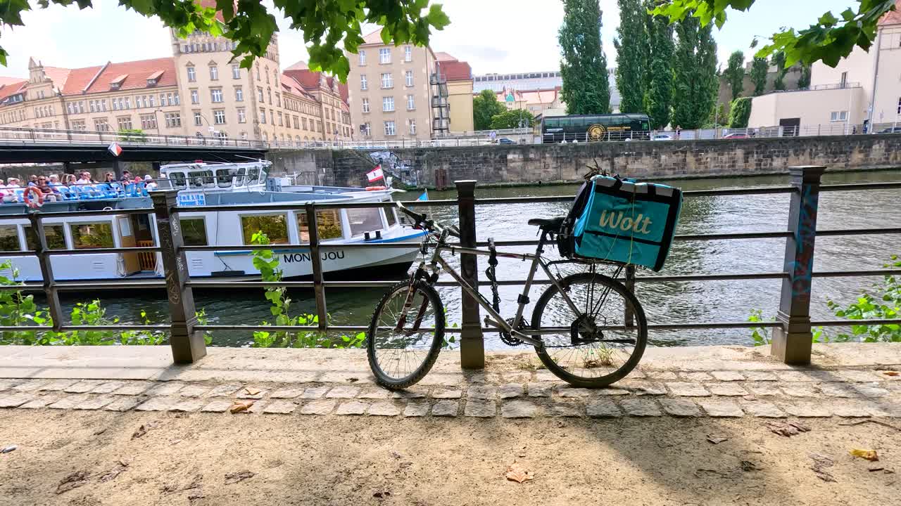 A sightseeing boat glides past a parked bicycle on a sunny day by the Spree River, with leafy trees and city buildings in the background