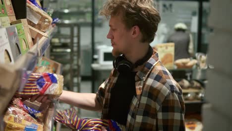 man in plaid shirt chooses a fresh bread loaf in the supermarket. young guy taking two loafs of bread from the shelf covered in bags. shopping in the grocery