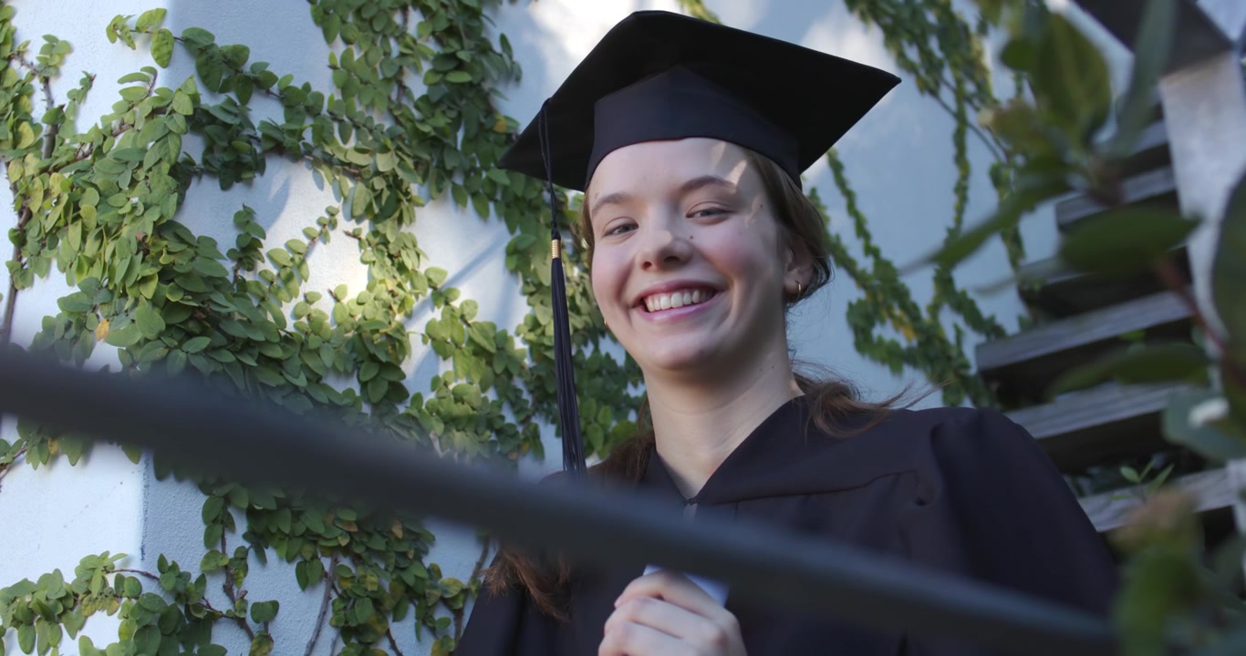 Donna laureata in toga e tocco sulla scalinata, sorride al segnale della telecamera alzando il mento e tenendo il diploma, festeggiando.