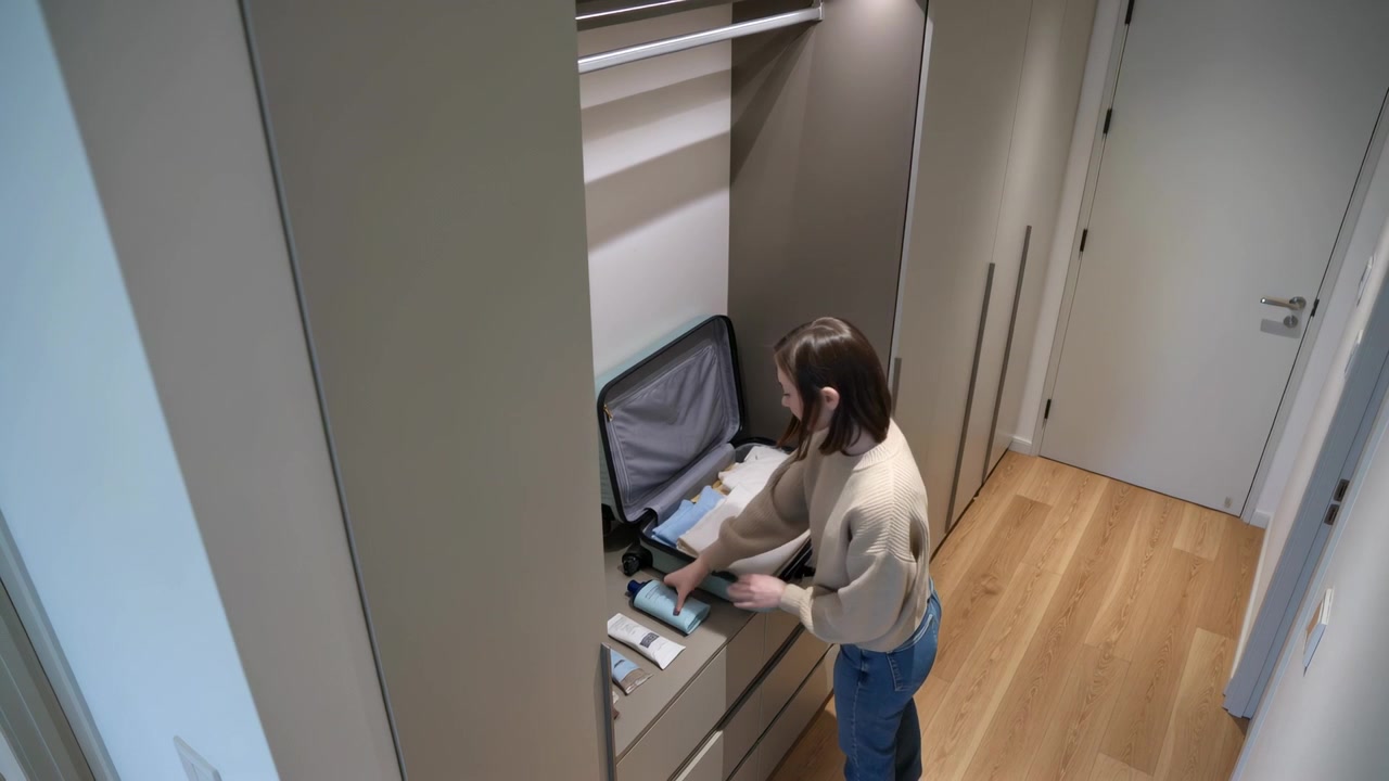 Donna con maglione leggero e jeans apre una valigia, prepara i vestiti e i bagagli in corridoio, con spazio per il testo.