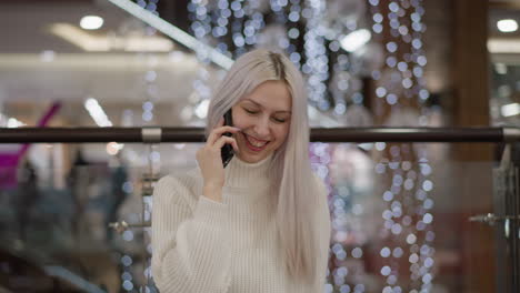 shoulder view of joyful lady on call, laughing heartily while seated on mall bench, wearing white sweater, jeans and boots, festive decorative lights and glass railing in background, retail interior