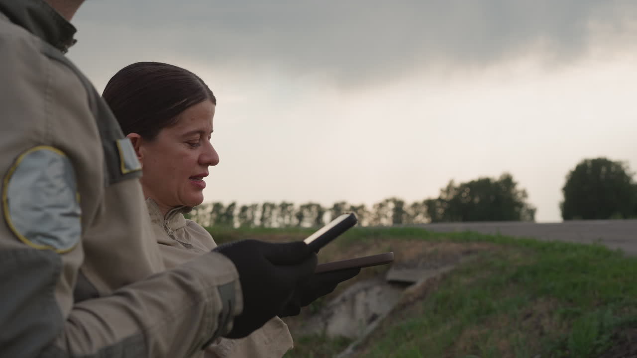 close up side view of two adults holding handheld devices monitoring roadside under overcast sky, wearing gloves and jackets, suggesting field survey and weather observation in rural environment