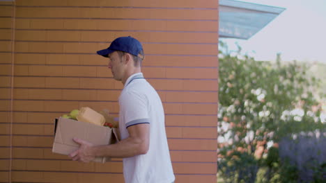 side view of male courier with blue cap coming to door of client with box of groceries and clipboard