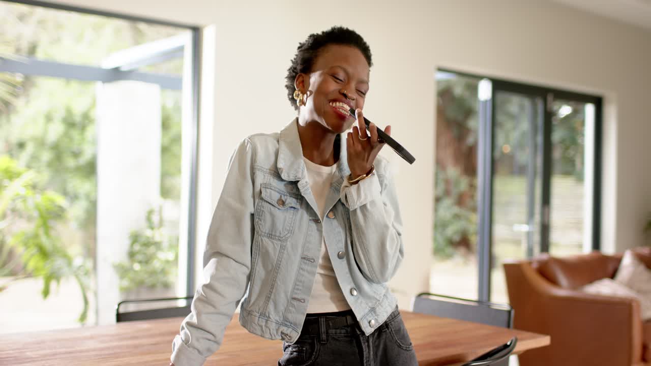 African American woman holding device like mic leaning at dining table at home practicing singing