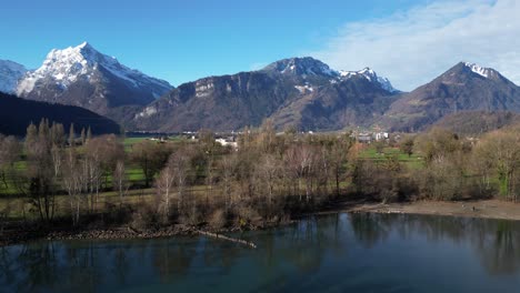 Clip-Panorámico-De-Drones-Que-Muestra-Montañas-Nevadas-En-Los-Alpes-Suizos,-Con-Tierras-Bajas-Cubiertas-De-Hierba-Y-La-Orilla-Del-Lago-En-Primer-Plano