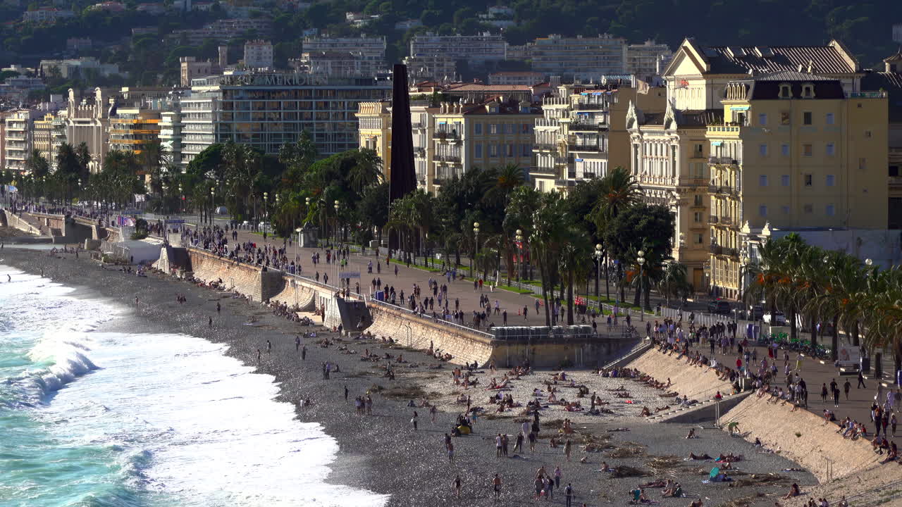 Turquoise waves crashing along the pebble beach of the Promenade des Anglais, with colorful buildings and palm trees lining the seafront
