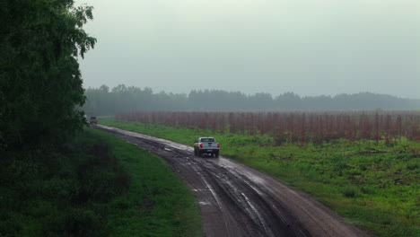 multiple vehicles on dirt road through forested farmland with green trees in hazy conditions